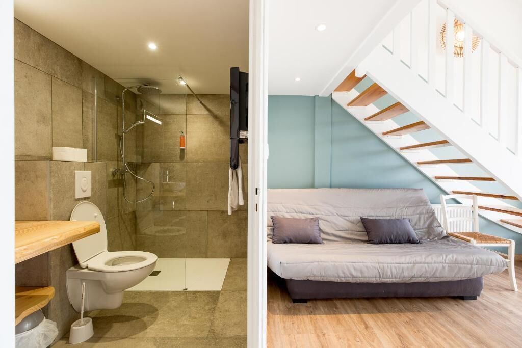 A modern bathroom with a shower, toilet, and sink. Adjacent is a bedroom with a bed and a staircase.