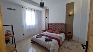 Um quarto com uma cama, mesa de cabeceira, luminária, janela com cortinas e piso de madeira.