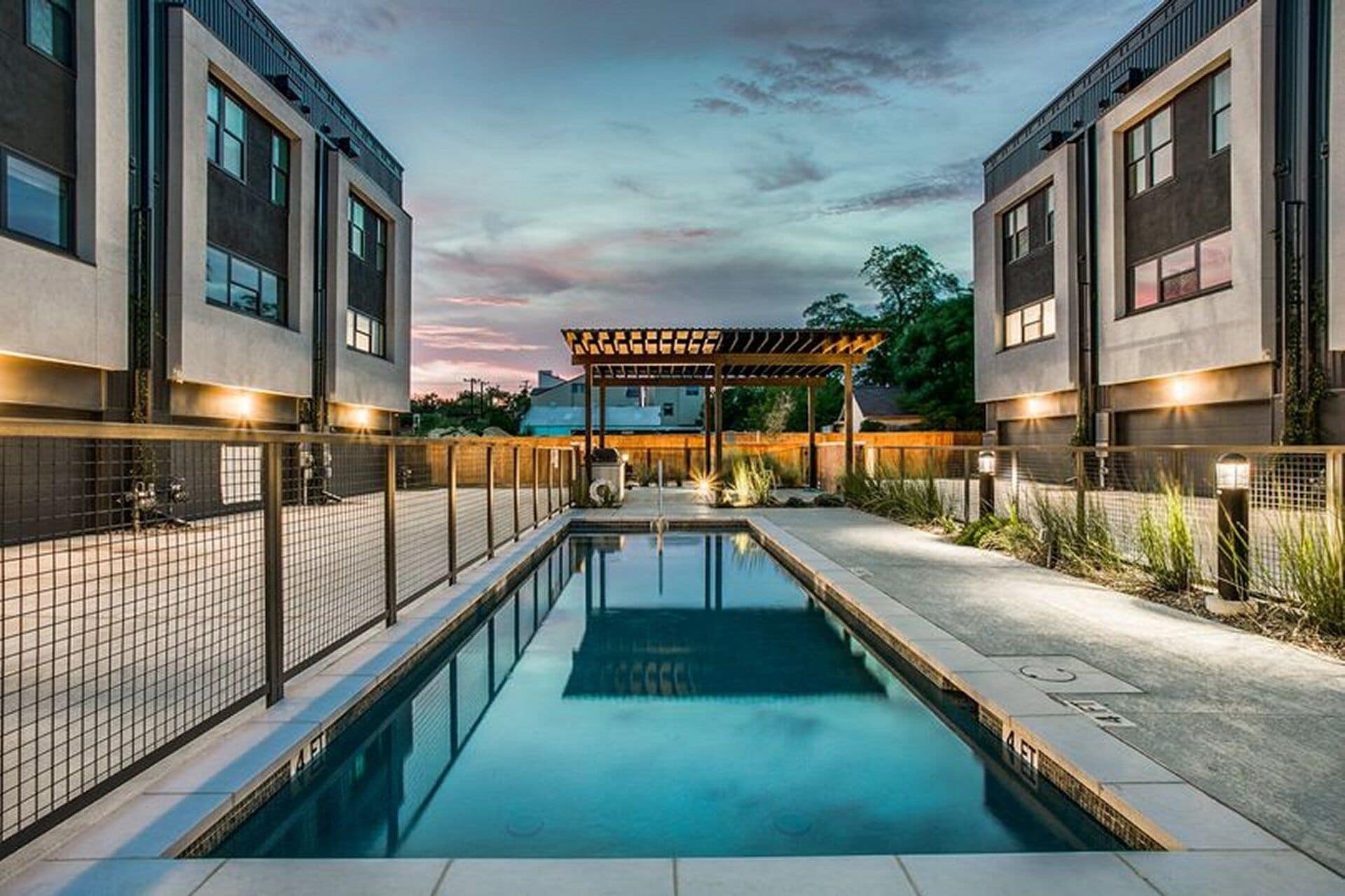 A modern pool area with a pergola, two-story buildings, and a clear sky.