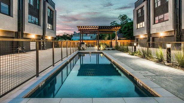 Un espace piscine moderne agrémenté d’une pergola, de bâtiments à deux étages et d’un ciel dégagé.