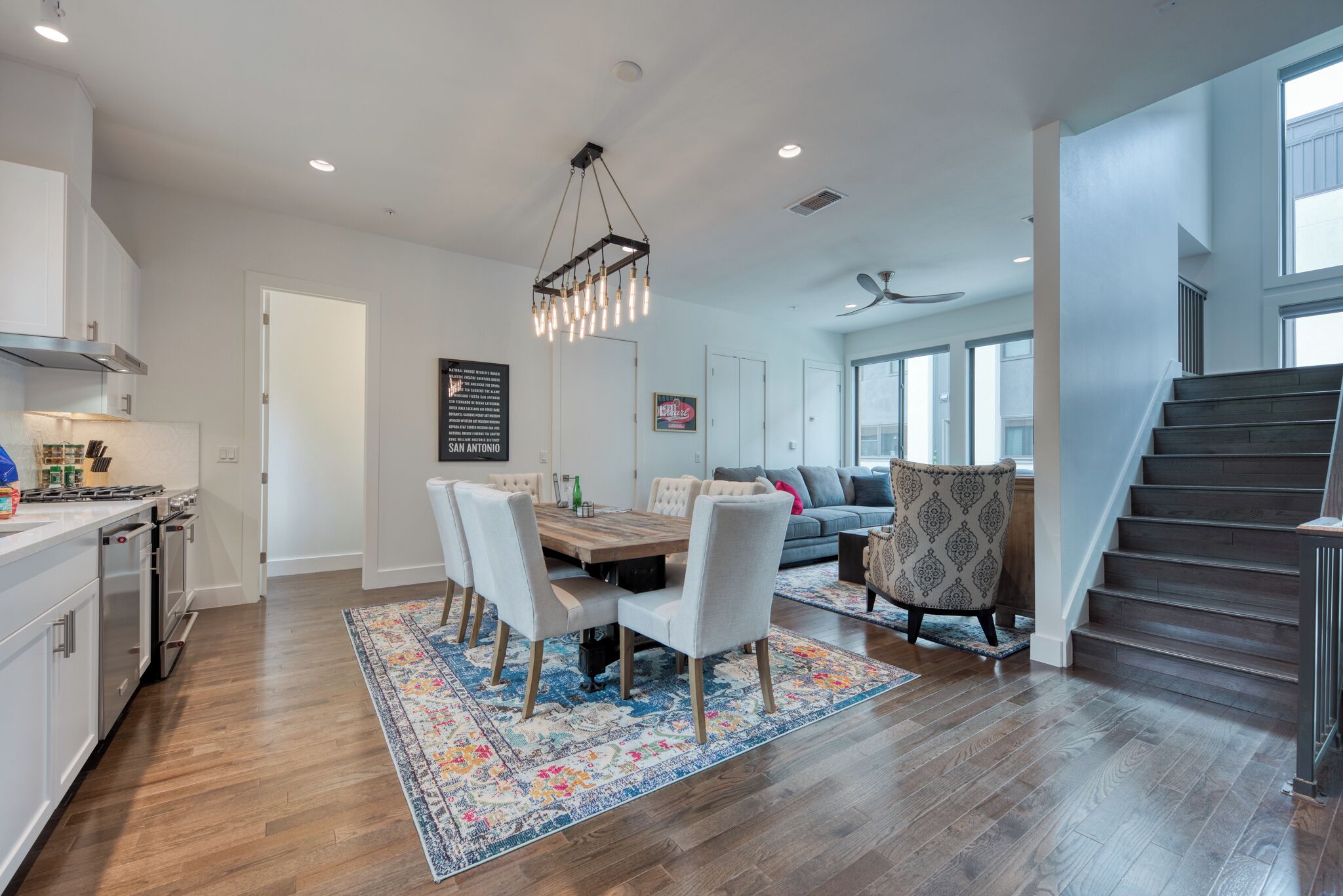 A modern kitchen with a dining area, a staircase, and a living room with a sofa and patterned armchair.