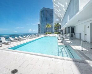 Ein Schwimmbad mit Liegestühlen, ein modernes Gebäude und der Blick aufs Meer.