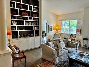 A living room with a large bookshelf, a sofa, a coffee table, and a window with shutters. - July in the Napa Valley (St. Helena)