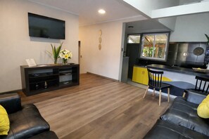 A modern living room with a flat-screen TV, a black entertainment unit, a dining area with a yellow cabinet, and a kitchen with a black countertop.