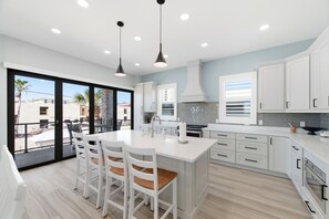 A modern kitchen with white cabinetry, a large island with bar stools, a balcony with a view, and a large window. - Strandhaus Unit 1: Luxury Retreat, Heated Pool, Elevator, Short Walk to Beach! (Indian Rocks Beach)