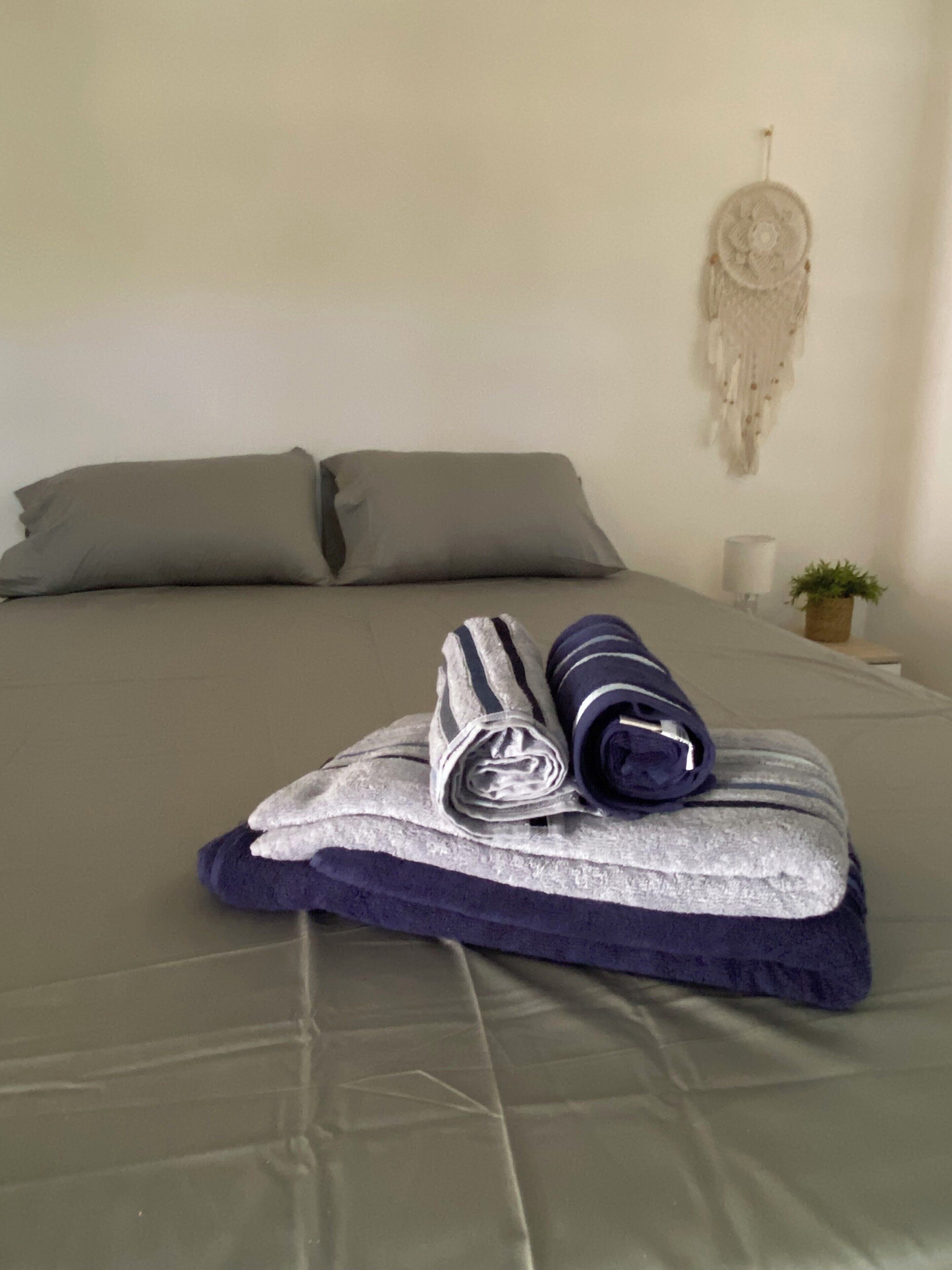 Een bed met een grijze lakenset, twee kussens, een opgevouwen handdoek, een dromenvanger, een lamp en een kleine plant.