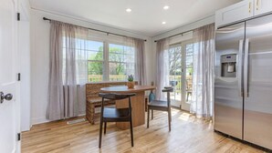 A kitchen with a stainless steel refrigerator, wooden flooring, a dining table with chairs, and large windows with curtains.