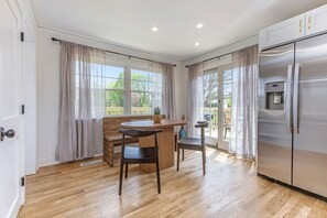 A kitchen with a stainless steel refrigerator, wooden flooring, a dining table with chairs, and large windows with curtains.