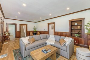 A living room with a sectional sofa, a wooden coffee table, a bookshelf, and a staircase.