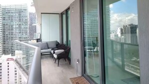 Un balcon avec un canapé et des fauteuils, de grandes portes vitrées et une vue sur les immeubles de la ville.