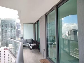 Ein Balkon mit einer Couch und Stühlen, großen Glasfenstern und Blick auf Stadtgebäude.