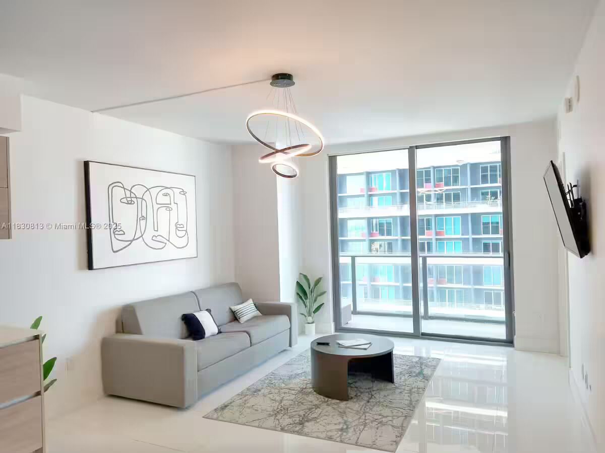 Un salon moderne avec un canapé gris, une table basse ronde, un lampadaire et une grande fenêtre donnant sur des immeubles.