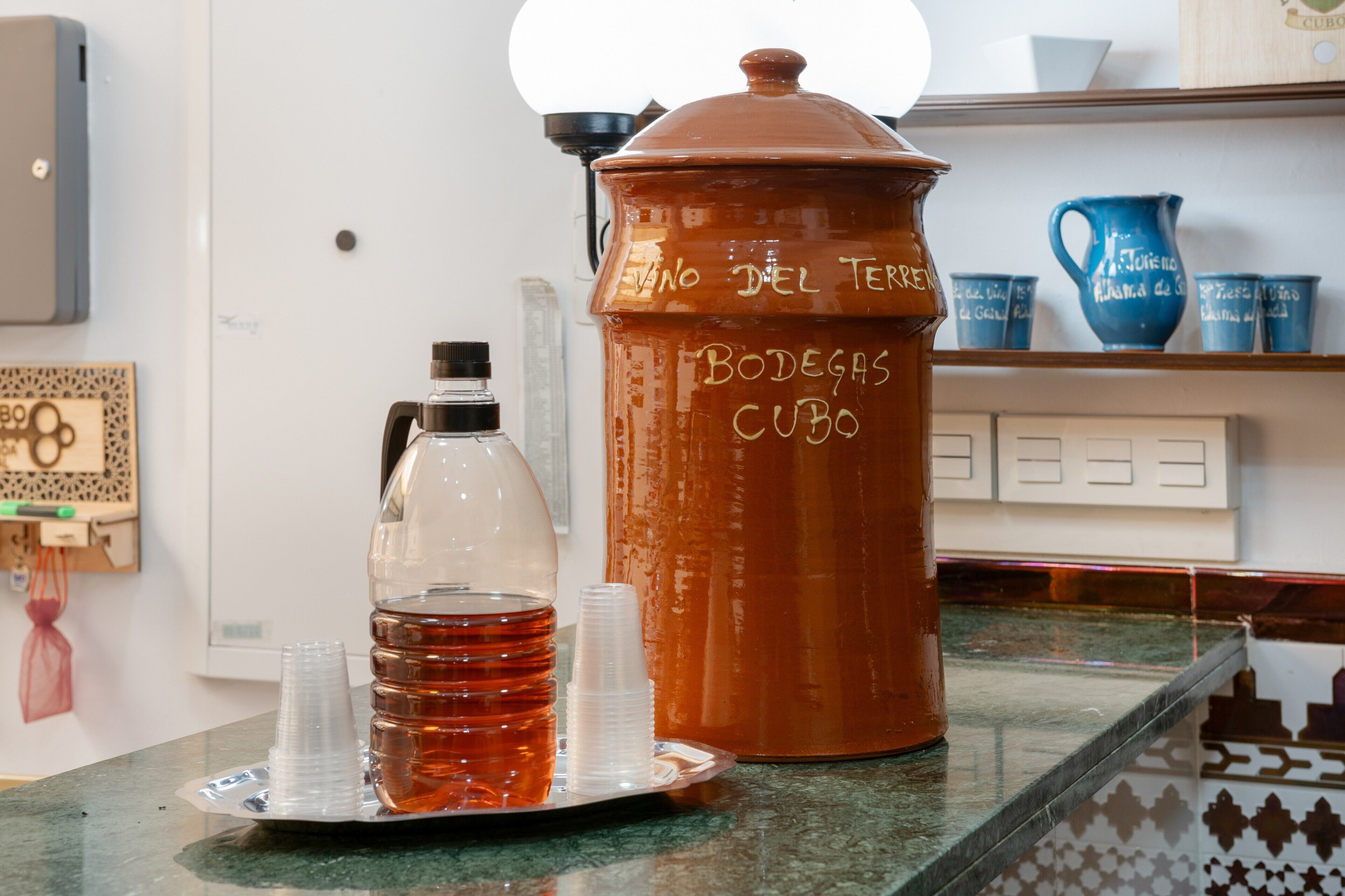 Un comptoir de cuisine sur lequel on trouve un grand pot en terre cuite étiqueté « Bodegas Cubo », une bouteille en plastique transparente et une pile de gobelets en plastique.
