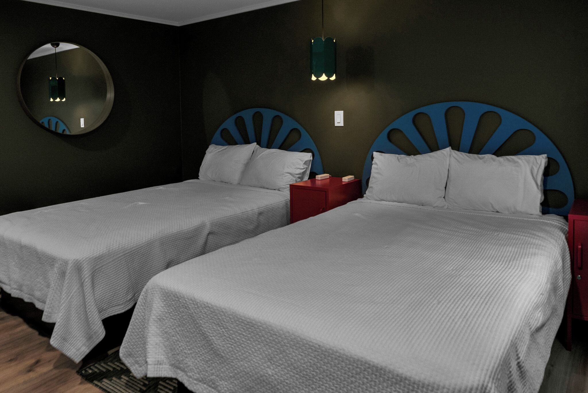 A room with two beds, each with a headboard featuring a blue fan-like design. There is a round mirror on the wall and a small table with a red lamp.