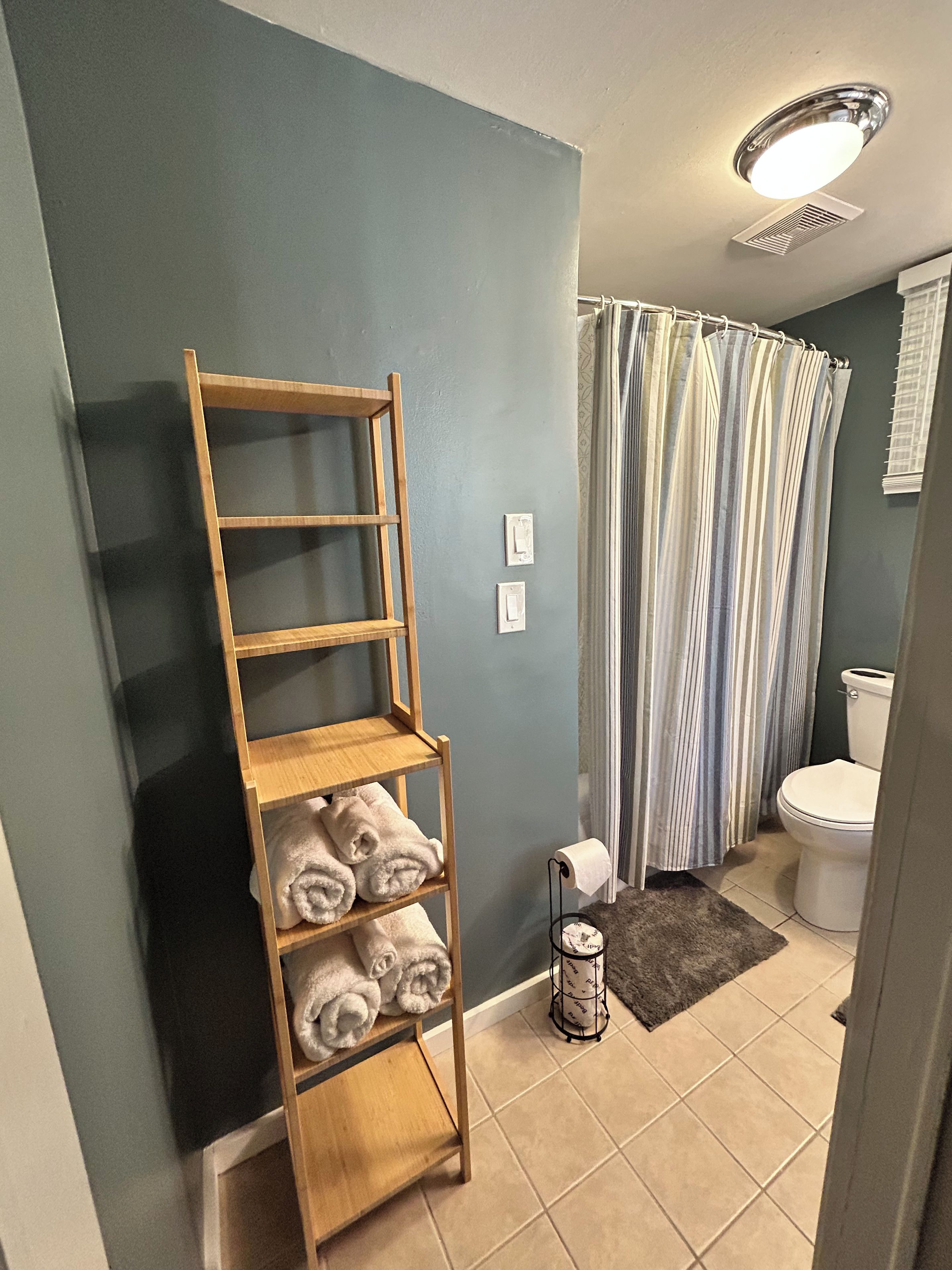 A bathroom with a wooden shelf holding towels, a striped shower curtain, a toilet, and a small rug on the floor.