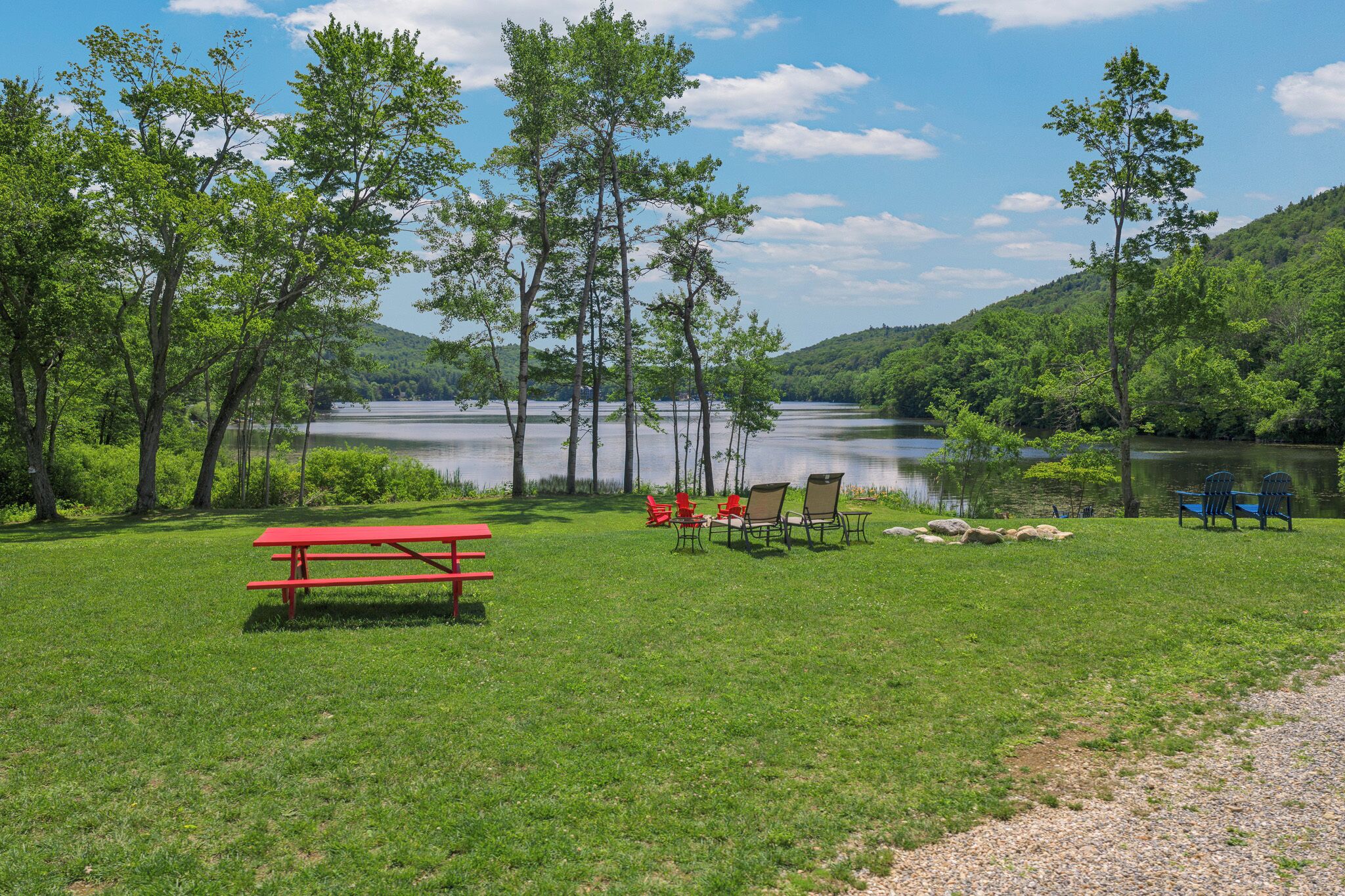 Zona de picnic junto al lago con una mesa roja de picnic, varias sillas Adirondack y vista a un lago rodeado de árboles.