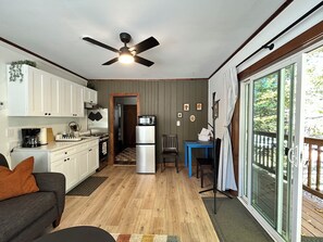 A compact kitchen with white cabinets, a refrigerator, and a microwave. There's a dining area with a blue table and chairs, and a large window offering a view of the outdoors. - Berkshire Lake Lodge  (Lee)