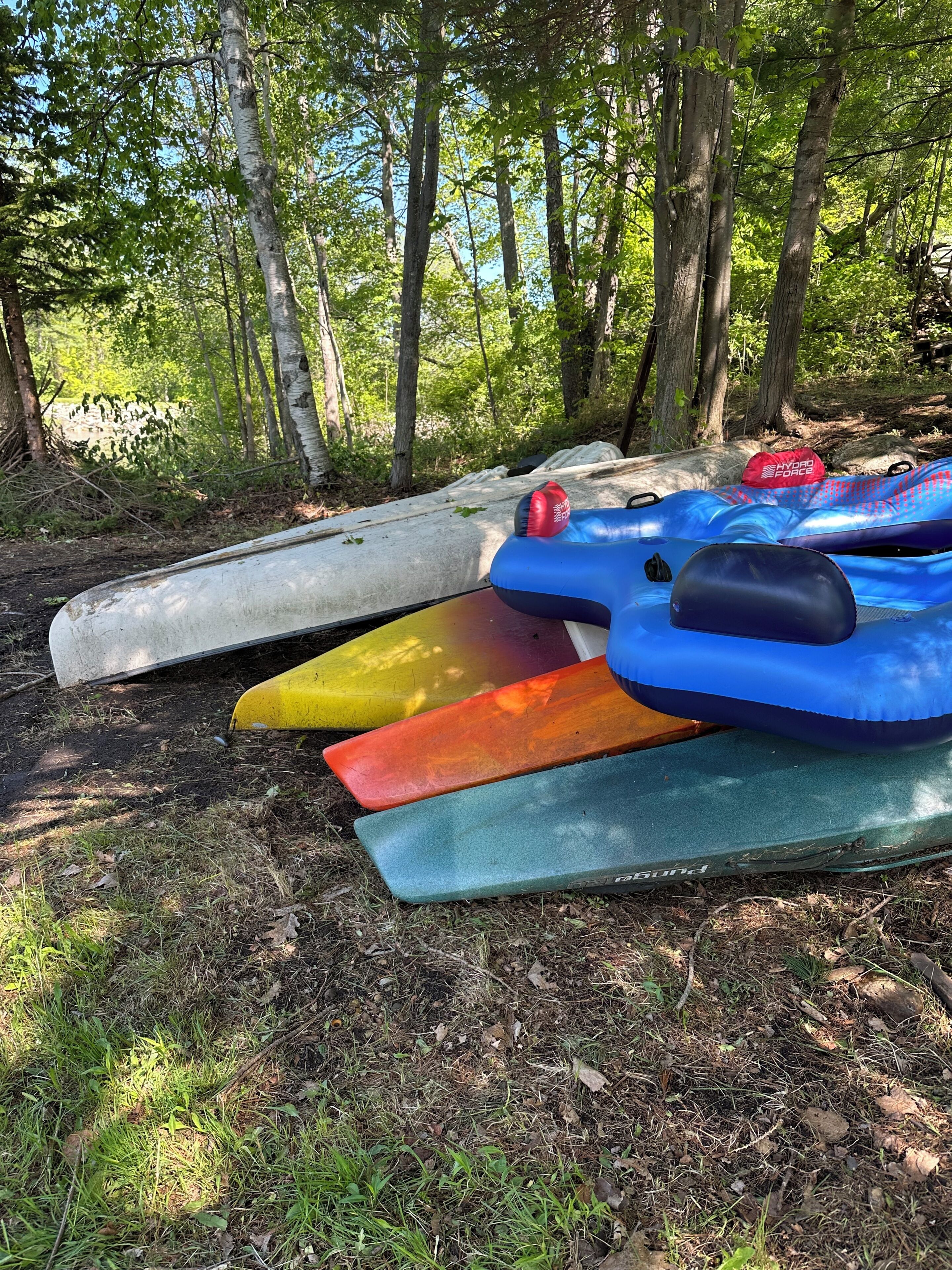A row of kayaks and a paddleboard resting on the ground in a wooded area.