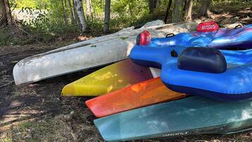 A row of kayaks and a paddleboard resting on the ground in a wooded area.