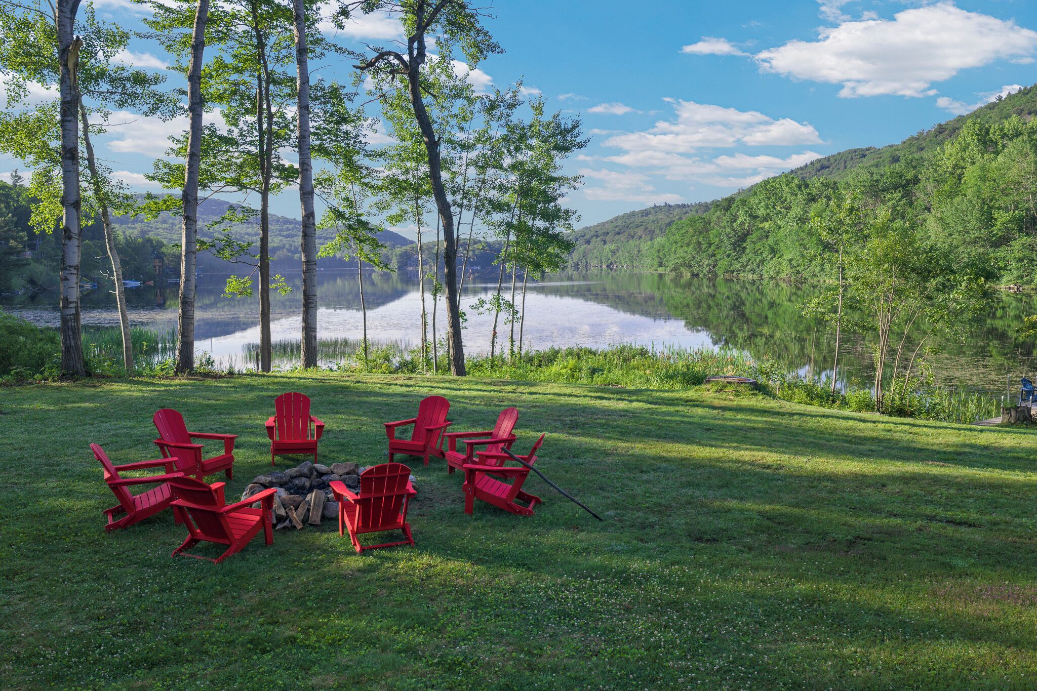 A grassy area with a lake, trees, and red chairs arranged around a fire pit.