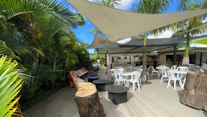 An outdoor seating area with tables and chairs, surrounded by palm trees and a shaded canopy. - La Casa 974 (Saint-Pierre)