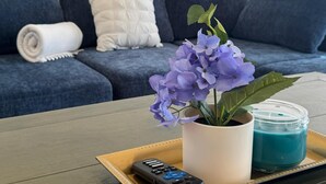 A living room with a blue sofa, a white cushion, a rolled white towel, a tray with a remote control, a potted plant, and a blue candle. - 2Bed1Bath Mostaccioli KingQueenBeds CentralPhoenix (Phoenix)