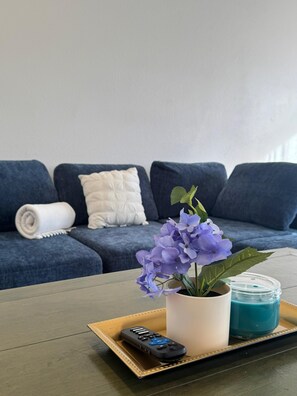 A living room with a blue sofa, a white cushion, a rolled white towel, a tray with a remote control, a potted plant, and a blue candle. - 2Bed1Bath Mostaccioli KingQueenBeds CentralPhoenix (Phoenix)