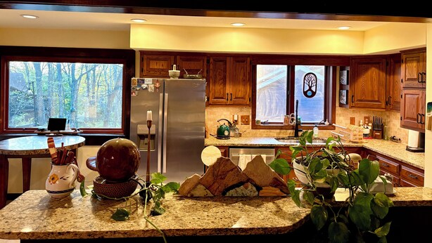 A kitchen with wooden cabinets, granite countertops, a refrigerator, and a window with a view of trees. - The Stone & Bridge: Close to MKT Trail & Dog Park, Dining, Groceries & I-70 (Columbia)