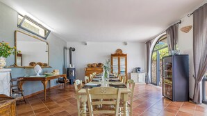 Ein Essbereich mit einem langen Tisch, gedeckt für sechs Personen, umgeben von Stühlen. Es gibt einen Holzschrank mit Glastüren, einen Spiegel an der Wand und ein Fenster mit Vorhängen.