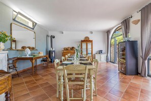 Un coin repas avec une longue table dressée pour six personnes, entourée de chaises. On y trouve un buffet en bois avec des portes en verre, un miroir au mur et une fenêtre avec des rideaux.