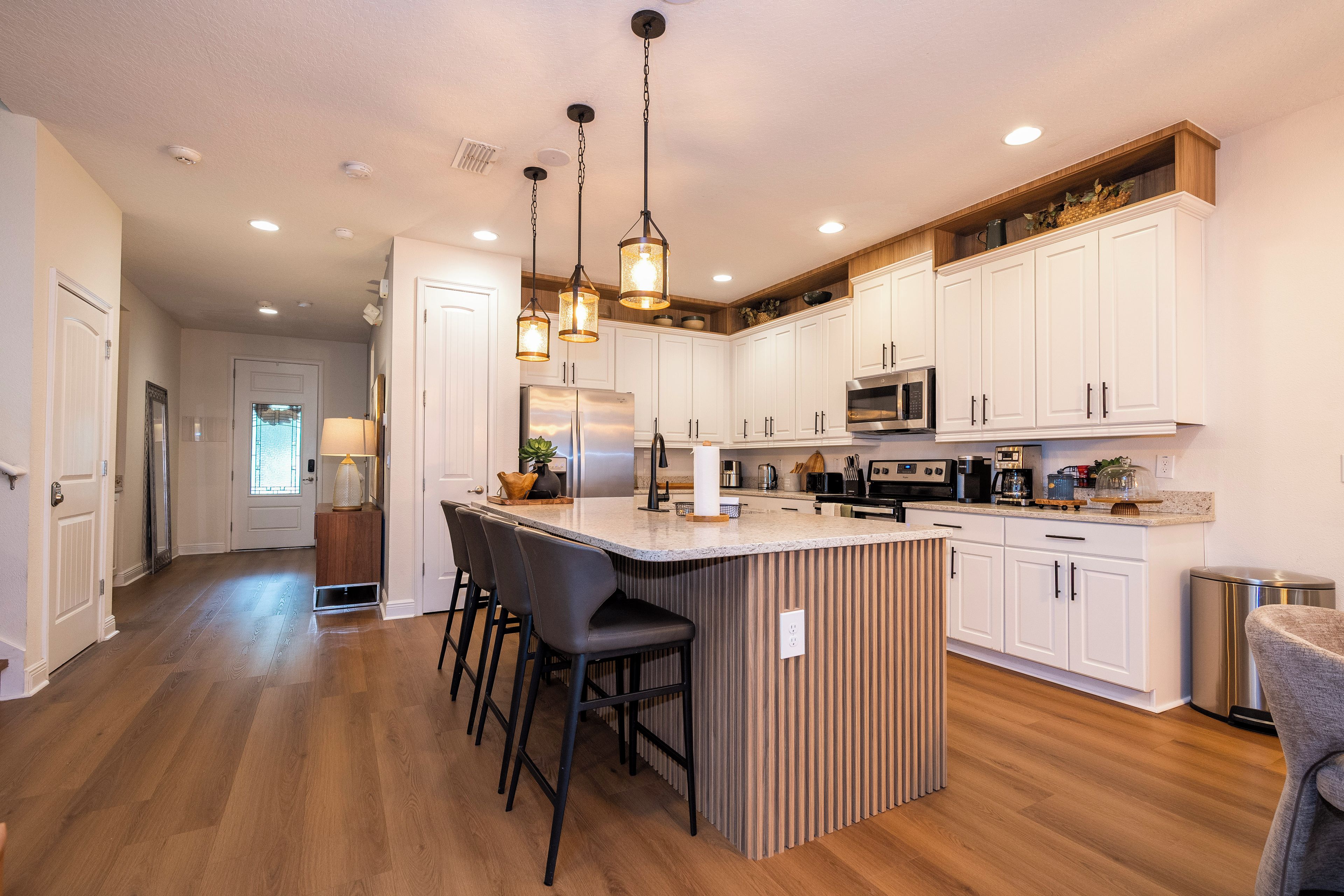 A modern kitchen with a central island, bar stools, white cabinets, stainless steel appliances, and pendant lighting.