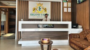Un mostrador de recepción con un cartel que dice 'Shahi Land Hotel', una araña, un televisor y una zona de recepción con un sofá y una mesita.