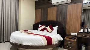 Una cama redonda con cabecera oscura, una colcha blanca y roja, y una mesita de noche de madera con un teléfono y un cubo de basura.