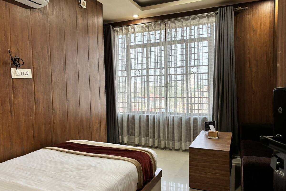 A hotel room with a bed, a desk, a chair, an air conditioner, and a window with curtains.