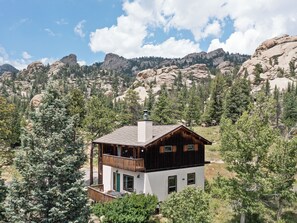 A two-story cabin with a balcony, surrounded by trees and mountains. - Thulite Chalet - 20-NCD0315 (Estes Park)