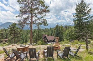 A rustic outdoor seating area with chairs and a round table, surrounded by tall pine trees and a mountain backdrop. - Thulite Chalet - 20-NCD0315 (Estes Park)
