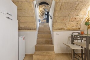 Eine enge Treppe führt zu einem Raum mit einer Steinwand, einem kleinen Tisch mit Stühlen und einem Kühlschrank.