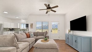 A modern living room with a sectional sofa, a ceiling fan, a flat-screen TV, and a kitchen in the background.