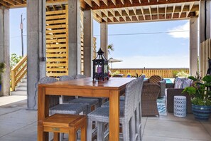 An outdoor patio with a wooden table and wicker chairs, a lantern, and a view of the beach. - New Luxury Beach Home - Heated Pool + Pickleball! (Pass Christian)