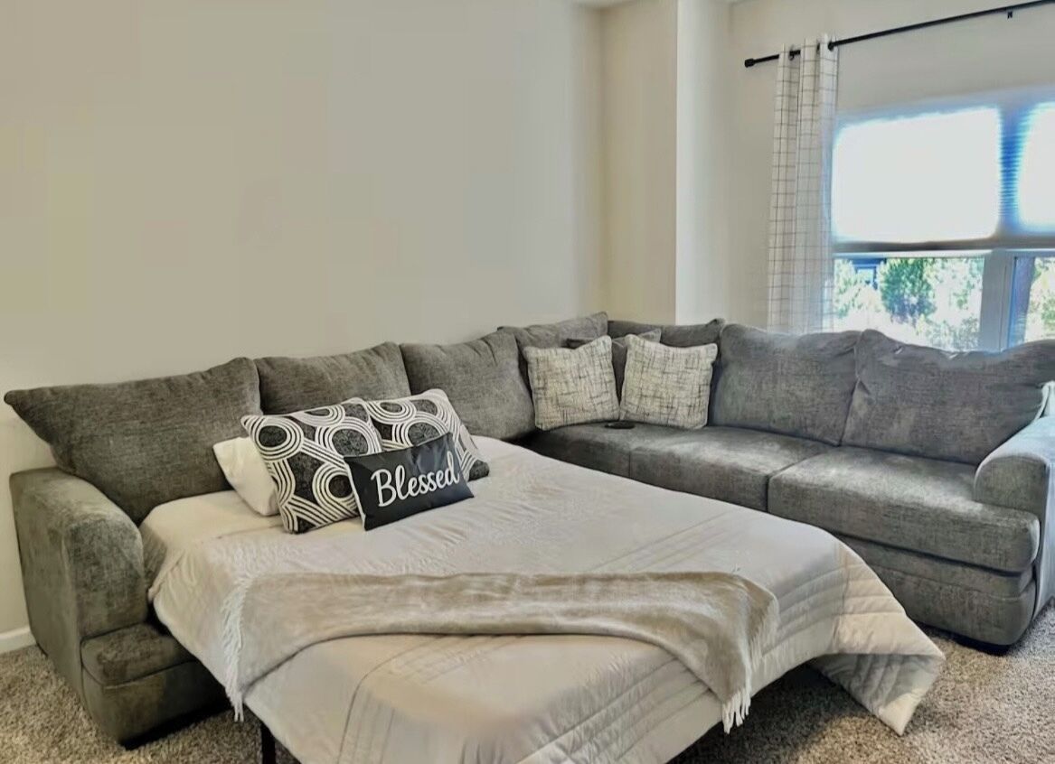A living room with a grey sectional sofa, a bed with a white comforter, and a window with blinds.