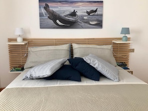 Un lit recouvert d’une couvre-lit à motifs, deux oreillers, un tableau représentant des bateaux accroché au mur, et des tables de chevet avec des lampes.