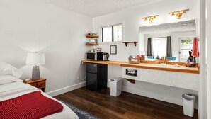 Una habitación compacta con cama, mesita de noche con lámpara, kitchenette con microondas, refrigerador y un pequeño bote de basura.