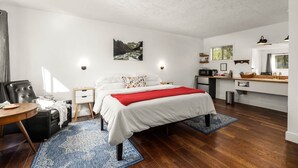 A bedroom with a bed, bedside tables, a desk, a chair, a small kitchen area, and a window. - Jacobson's Cottages Cabin # 2 (East Glacier Park)