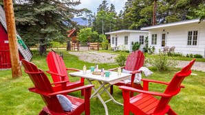 A cozy outdoor seating area with red Adirondack chairs and a table, surrounded by a lush green lawn and a white building in the background. - Jacobson's Cottages Cabin #7 (East Glacier Park)