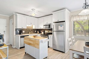 A modern kitchen with white cabinets, a central island, stainless steel appliances, and a dining area with a table and chairs. - 2BR Beach Villa with Balcony Pool Tennis Access (Hilton Head Island)
