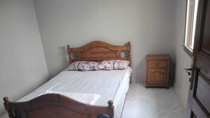 Ein Einzelbett mit Holz-Kopfstück, ein kleiner Nachttisch und eine schlichte Wand mit Fenster.