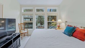 Een slaapkamer met een bed, een televisie, een stoel, een nachtkastje met een lamp en een raam met uitzicht op bomen en gebouwen.