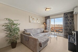A living room with a sofa, a potted plant, a painting, and a view of buildings through a window. - Vacation Apartment 'Vista Tejita' with Sea View, Wi-Fi and Air Conditioning (Granadilla)