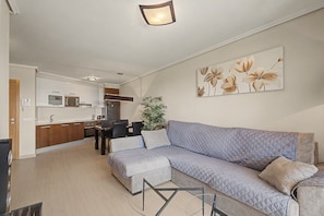 A modern living room with a sectional sofa, a dining area with chairs, a kitchen with appliances, and a large floral artwork on the wall.