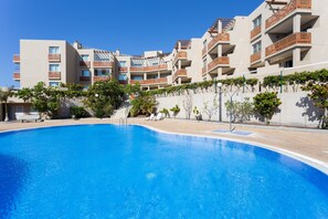 Une piscine à l’eau d’un bleu limpide, entourée d’un bâtiment doté de nombreux balcons et d’un jardin planté d’arbres et de fleurs.